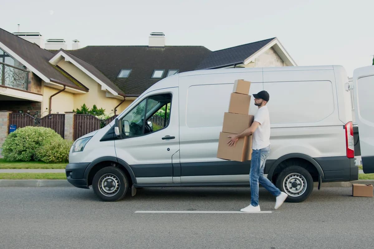A man carrying a stack of cardboard boxes toward an open van parked on a residential street.
