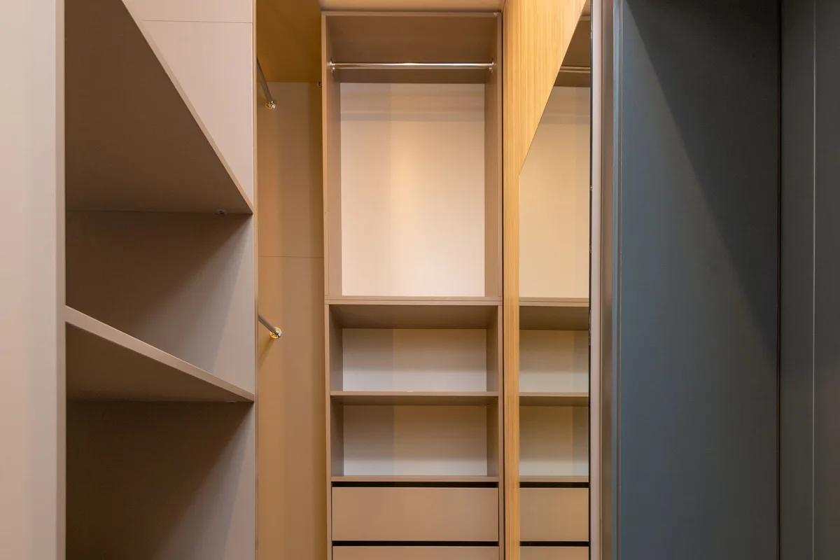 A built-in wardrobe with emptied shelves, drawers and a hanging rail, fitted into an alcove with a wooden floor.