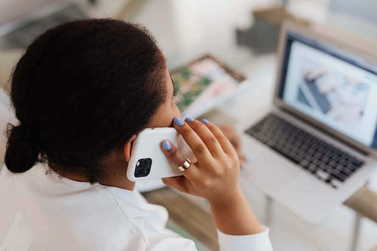 A woman holding a phone to her ear while looking at a laptop screen on a white table at home.