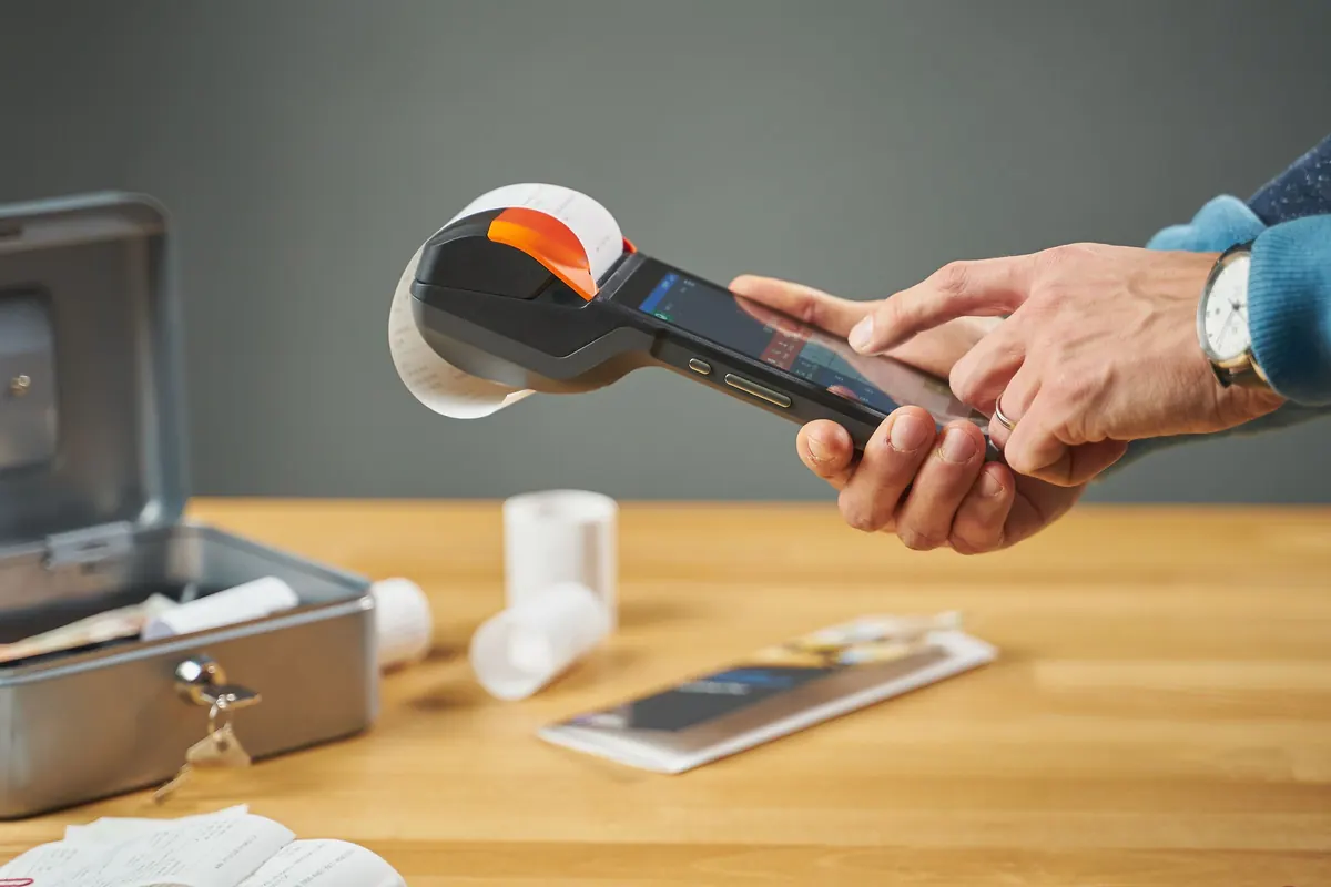 A handheld card terminal printing a paper receipt, held above a table with a cash box and receipt rolls.