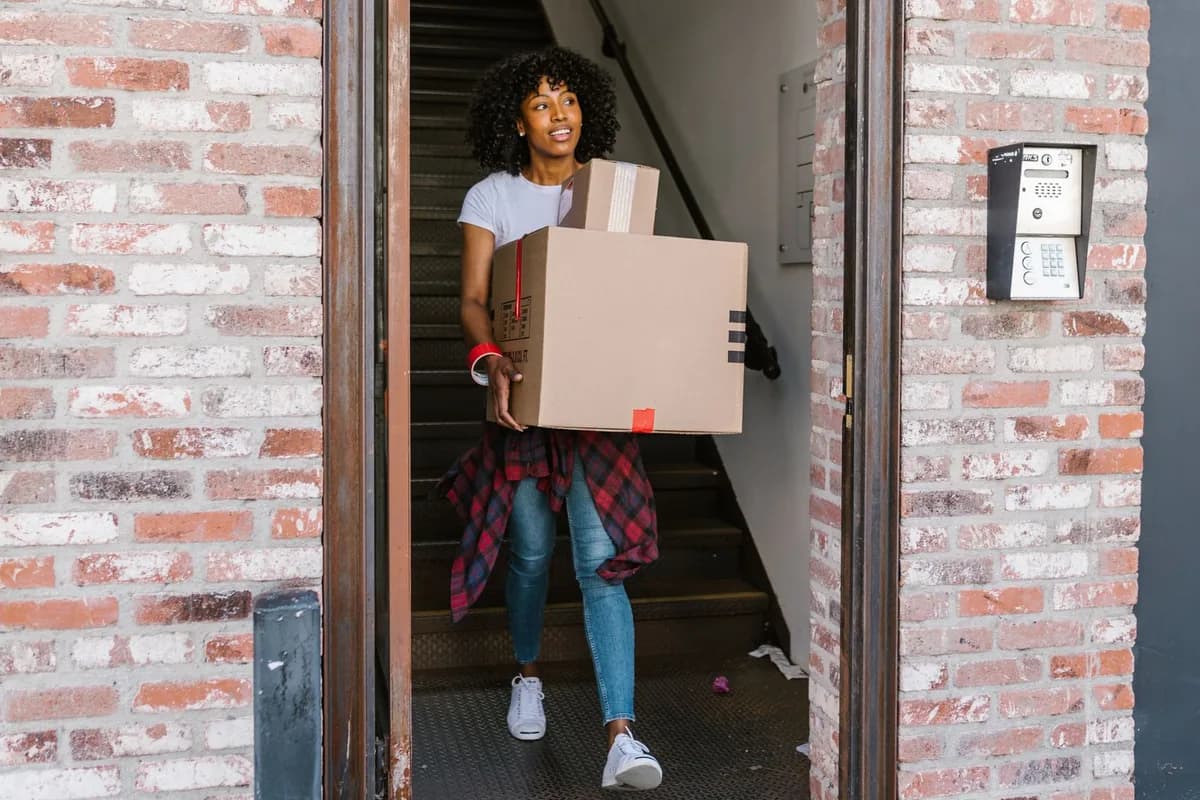 A woman carrying two cardboard boxes down an internal stairwell and out through a building's street entrance.