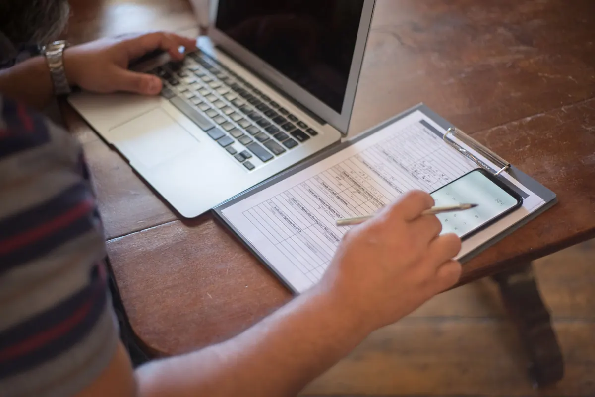 A person filling in a printed form on a clipboard beside an open laptop and a phone on a wooden table.