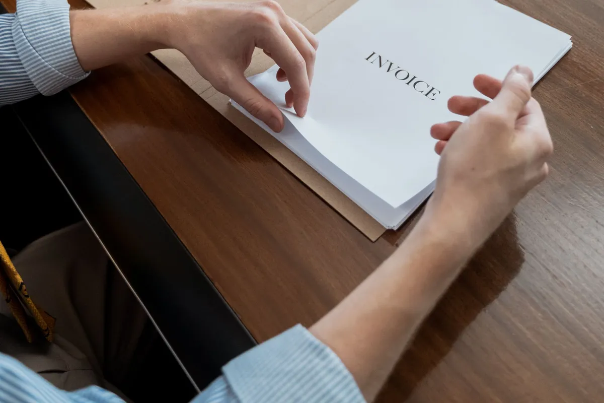 A person turning the pages of a stapled paper invoice on a polished wooden desk.