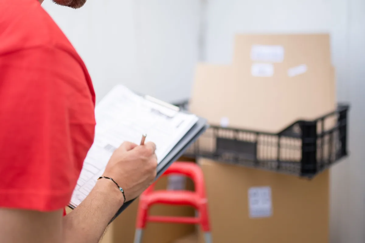A worker checking a written list against a stack of cardboard boxes and a crate.