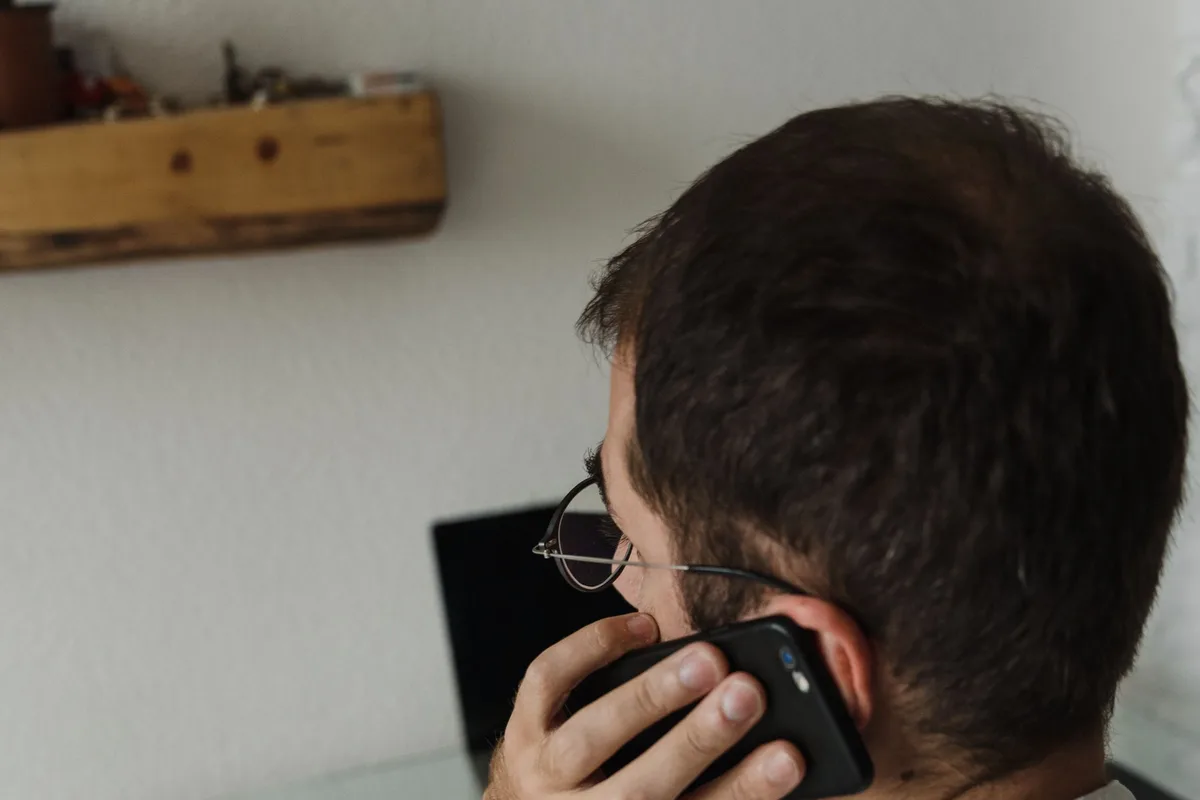 A man in a white shirt holding a phone to his ear at home, mid conversation.