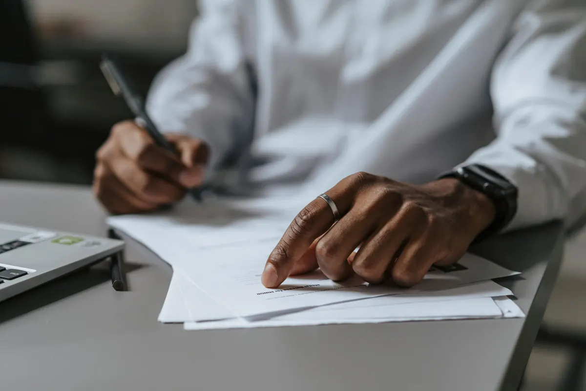 A person in a white shirt writing on a printed page with a pen, with a laptop open beside them on a desk.
