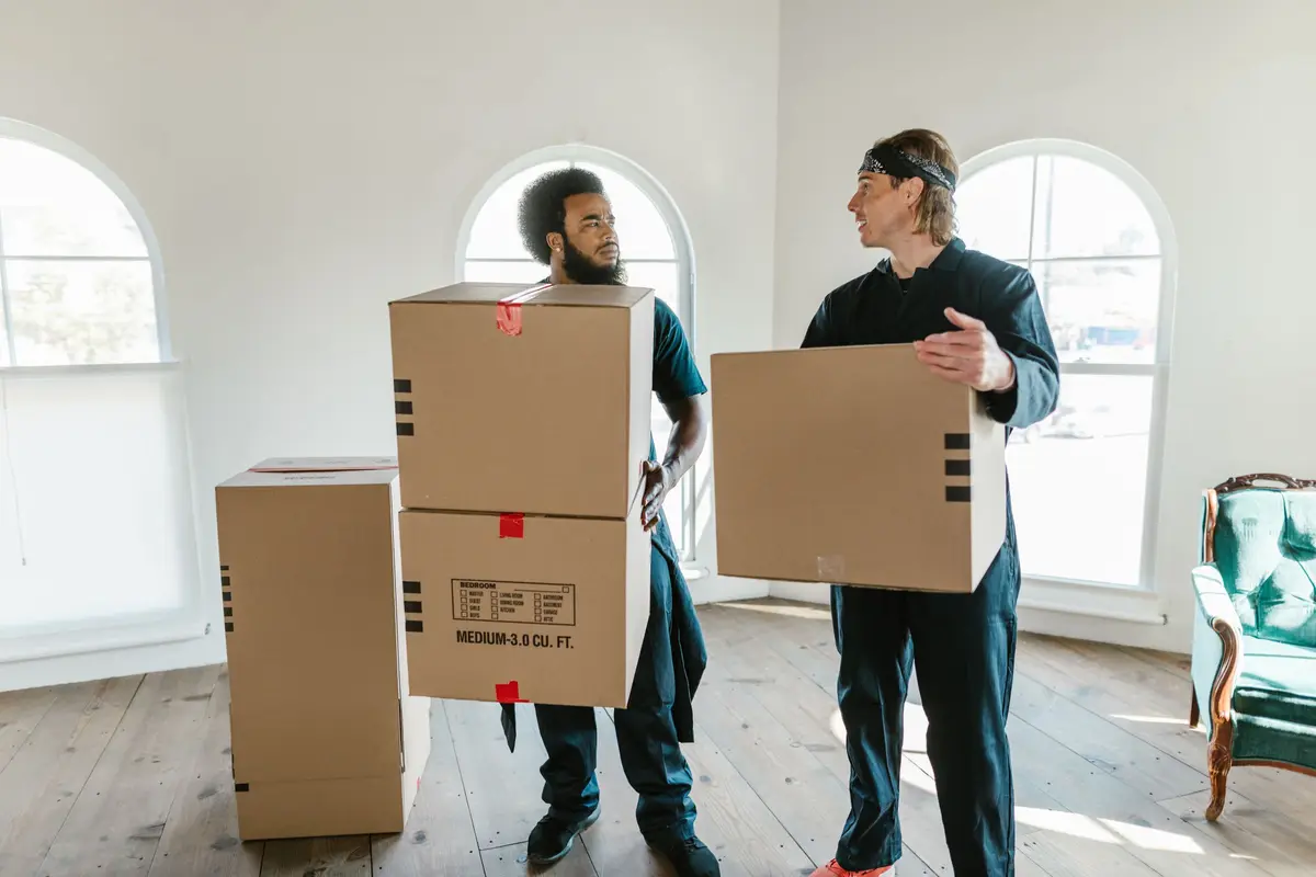 Two workers in dark overalls carrying stacked cardboard boxes out of an empty room with tall arched windows.