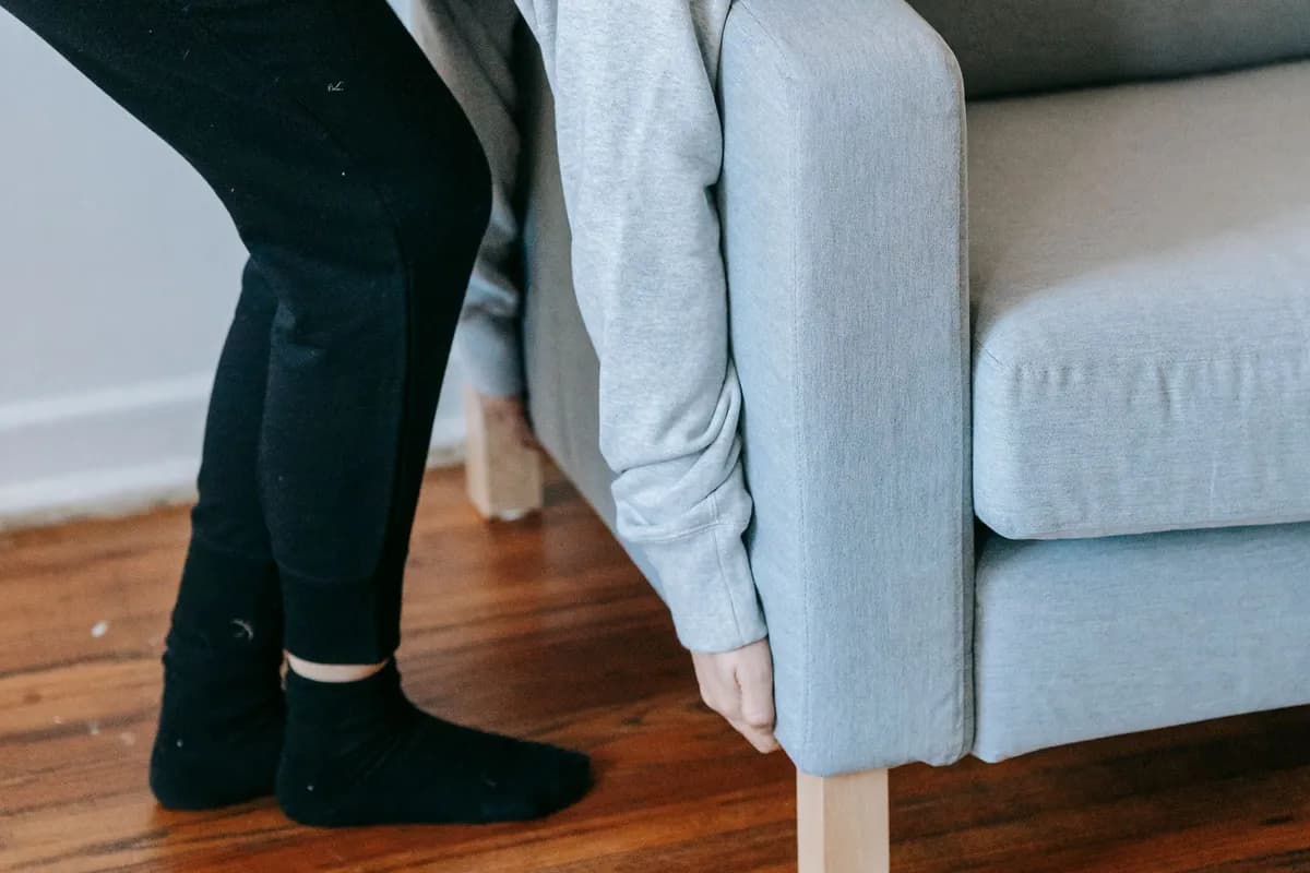 A person bracing on a wooden floor while gripping the underside of a sofa arm to lift one end on their own.
