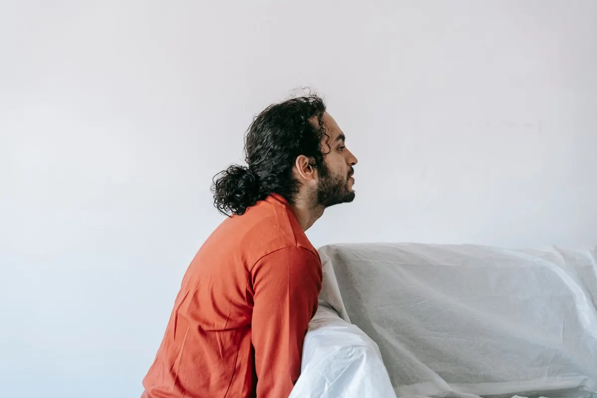 A man in an orange shirt lifting one end of a plastic-wrapped sofa in an otherwise empty room.