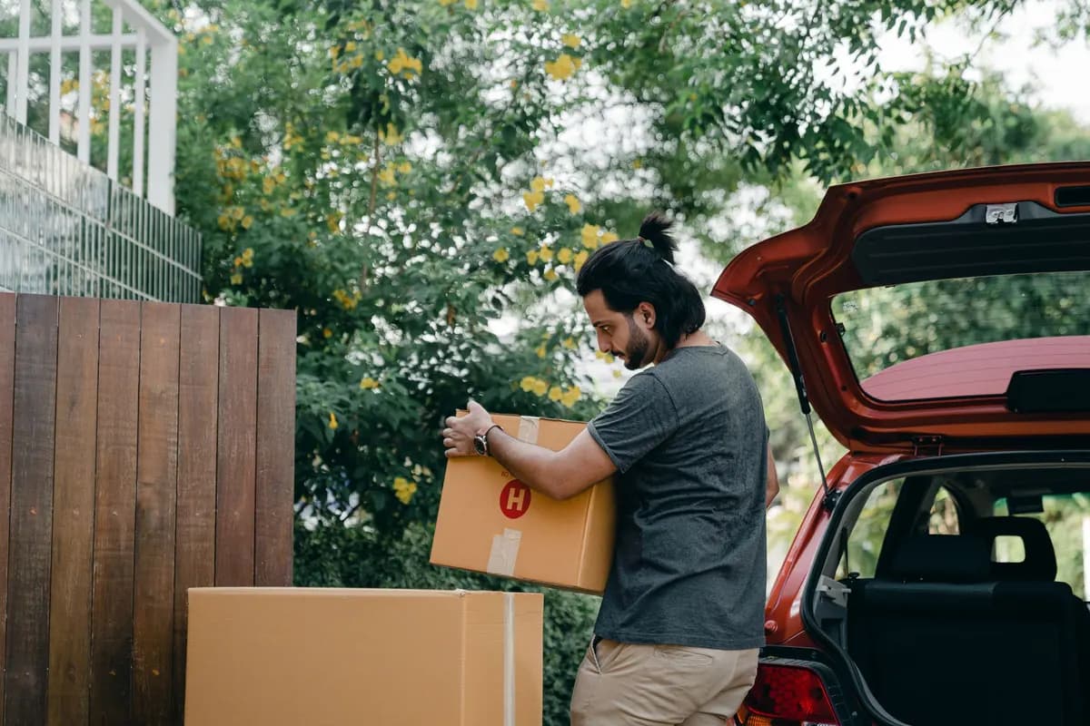 A man lifting a large taped cardboard box into the open boot of a car parked beside a garden wall.
