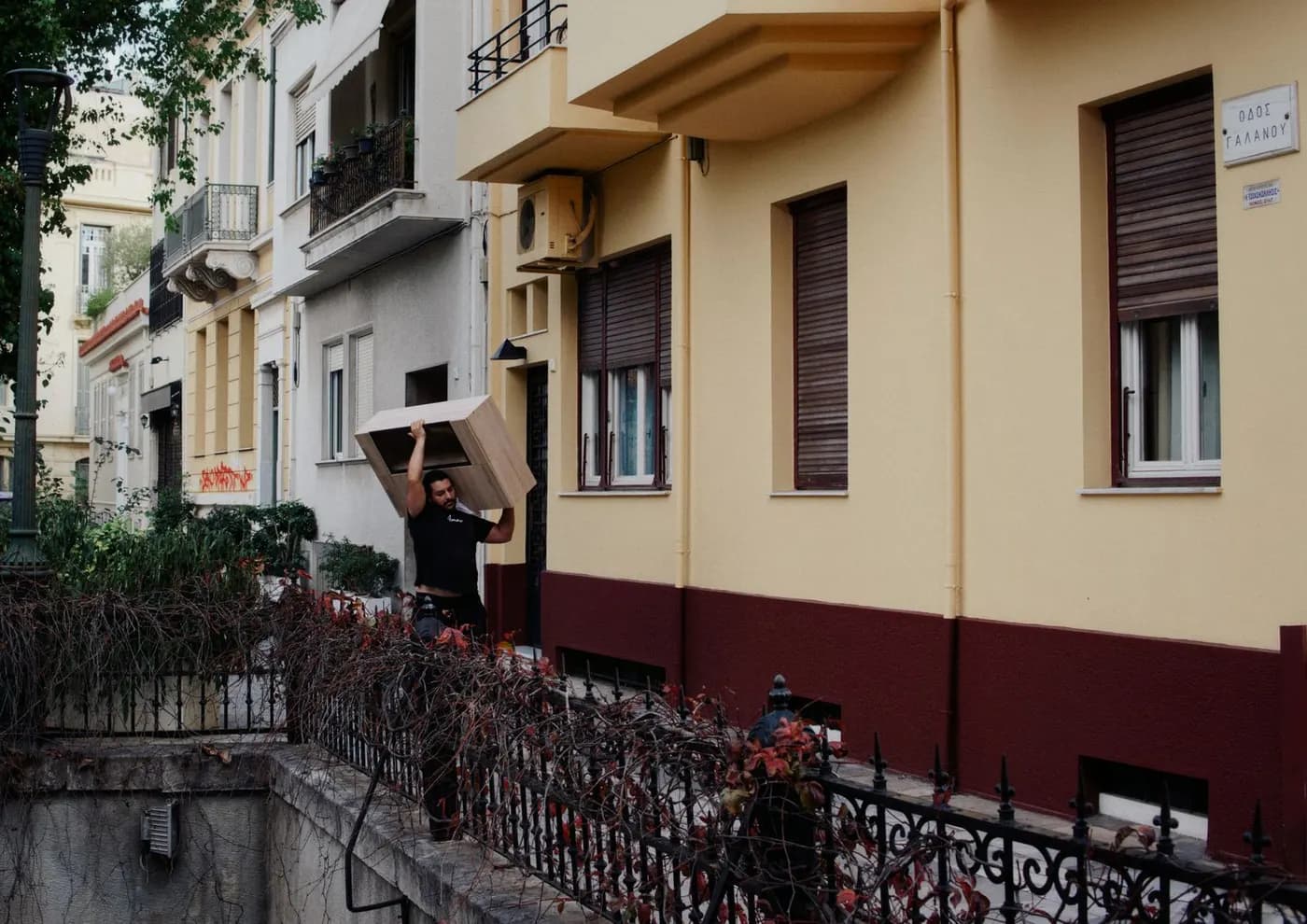 A man carrying a wooden cabinet balanced on his shoulder along a residential street lined with apartment buildings.