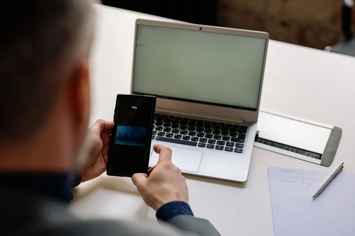 A person holding a phone showing a payment screen, with a laptop and a handwritten note on the desk beside them.
