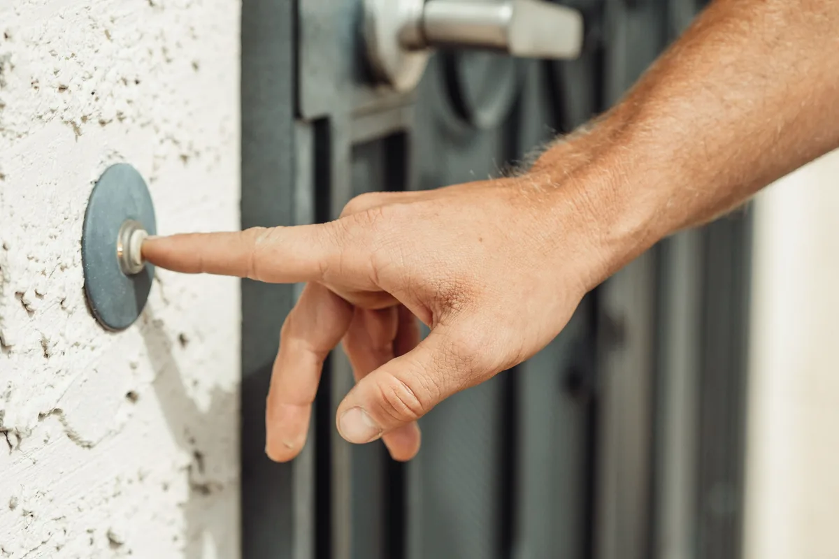 A hand pressing a doorbell button mounted beside a gated entrance.