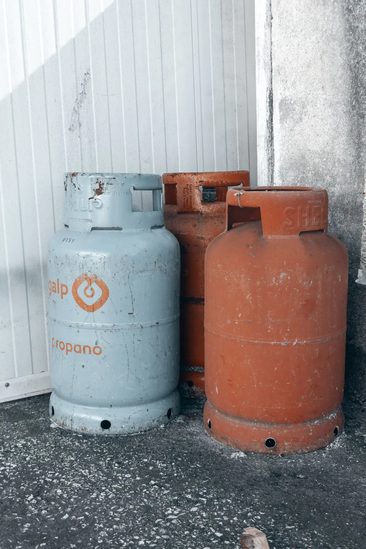 Three worn propane gas cylinders in different colours standing against a concrete wall.
