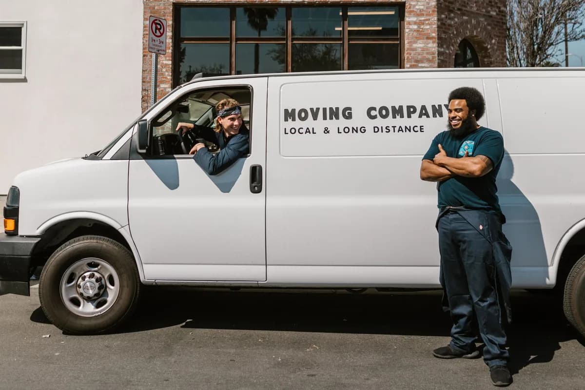 A white moving van parked at the kerb outside a two-storey building, with two crew members beside it and a no-parking sign on the pavement.