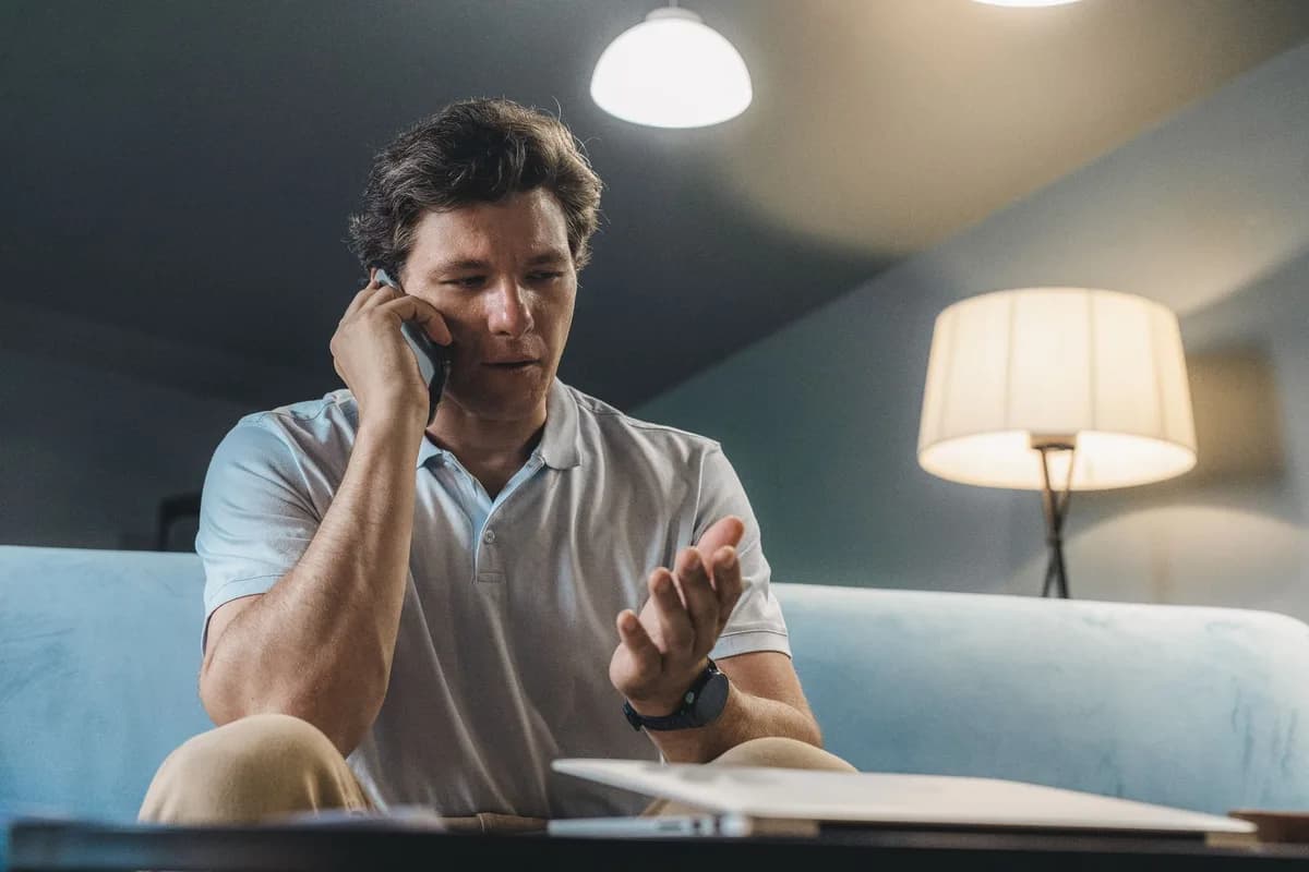 A man sitting on a sofa at home speaking on a mobile phone with papers open on the table in front of him.