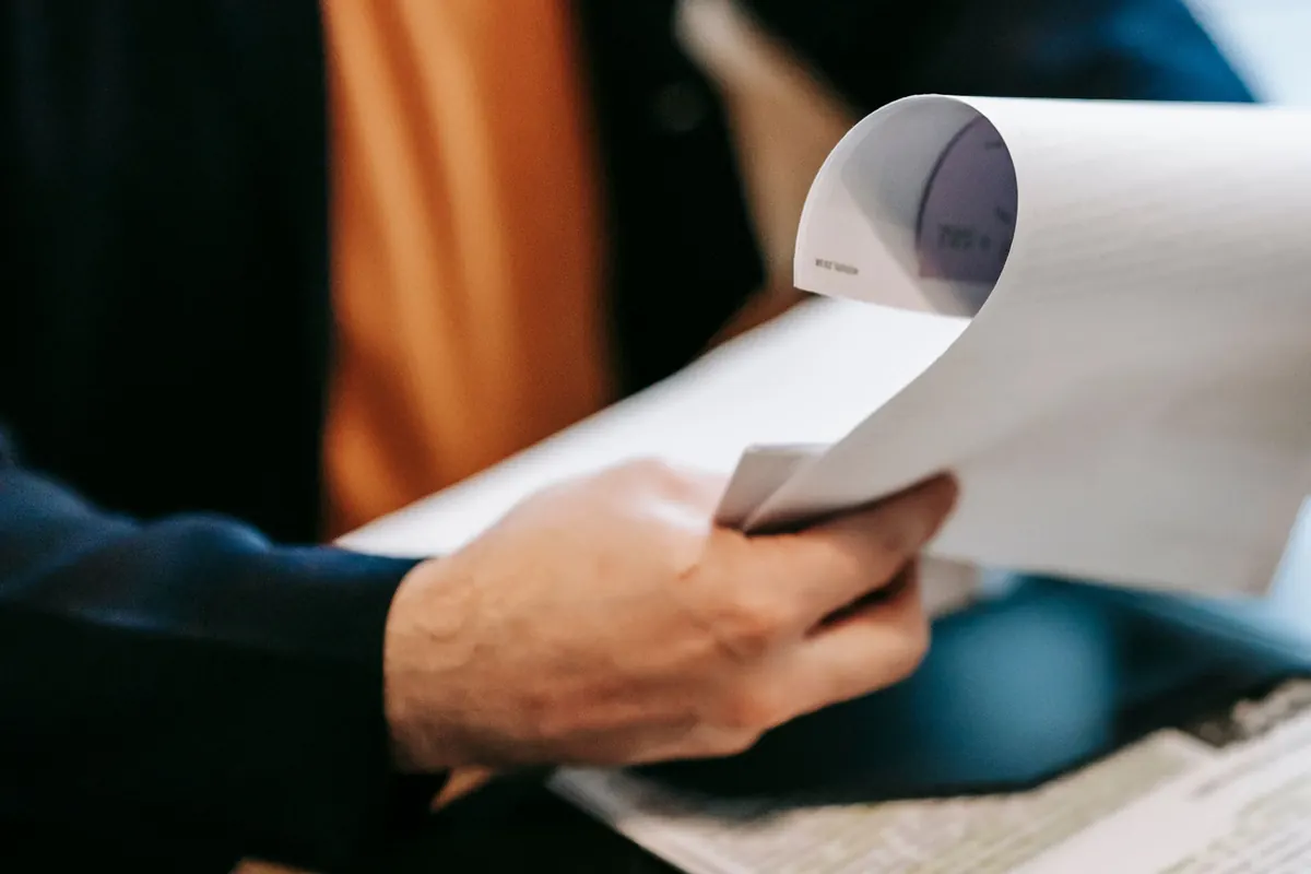 A person in a dark jacket turning the page of a printed document held in one hand.