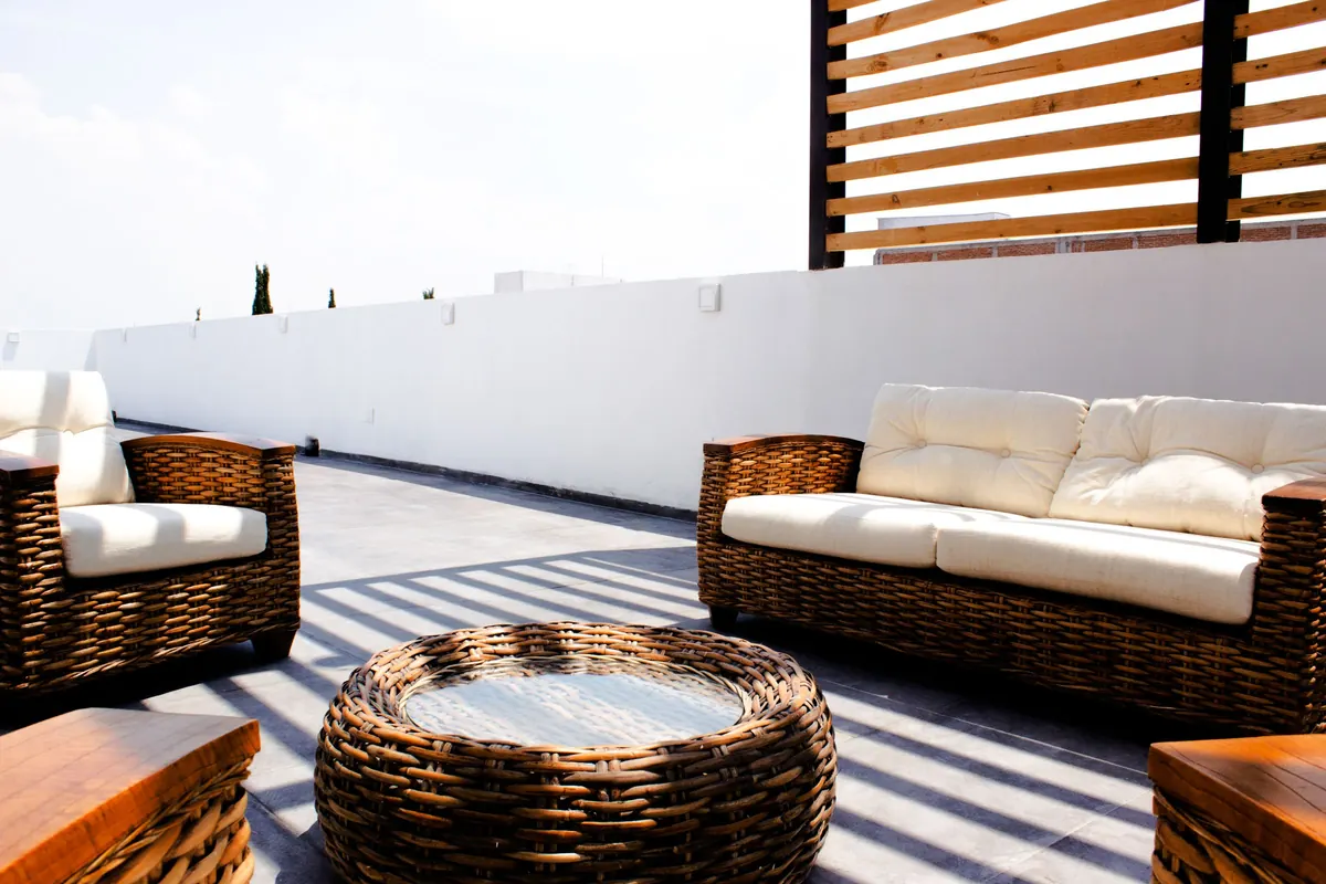 Rattan sofas and a round wicker table on a sunlit roof terrace with a white parapet wall and a timber slatted shade screen.