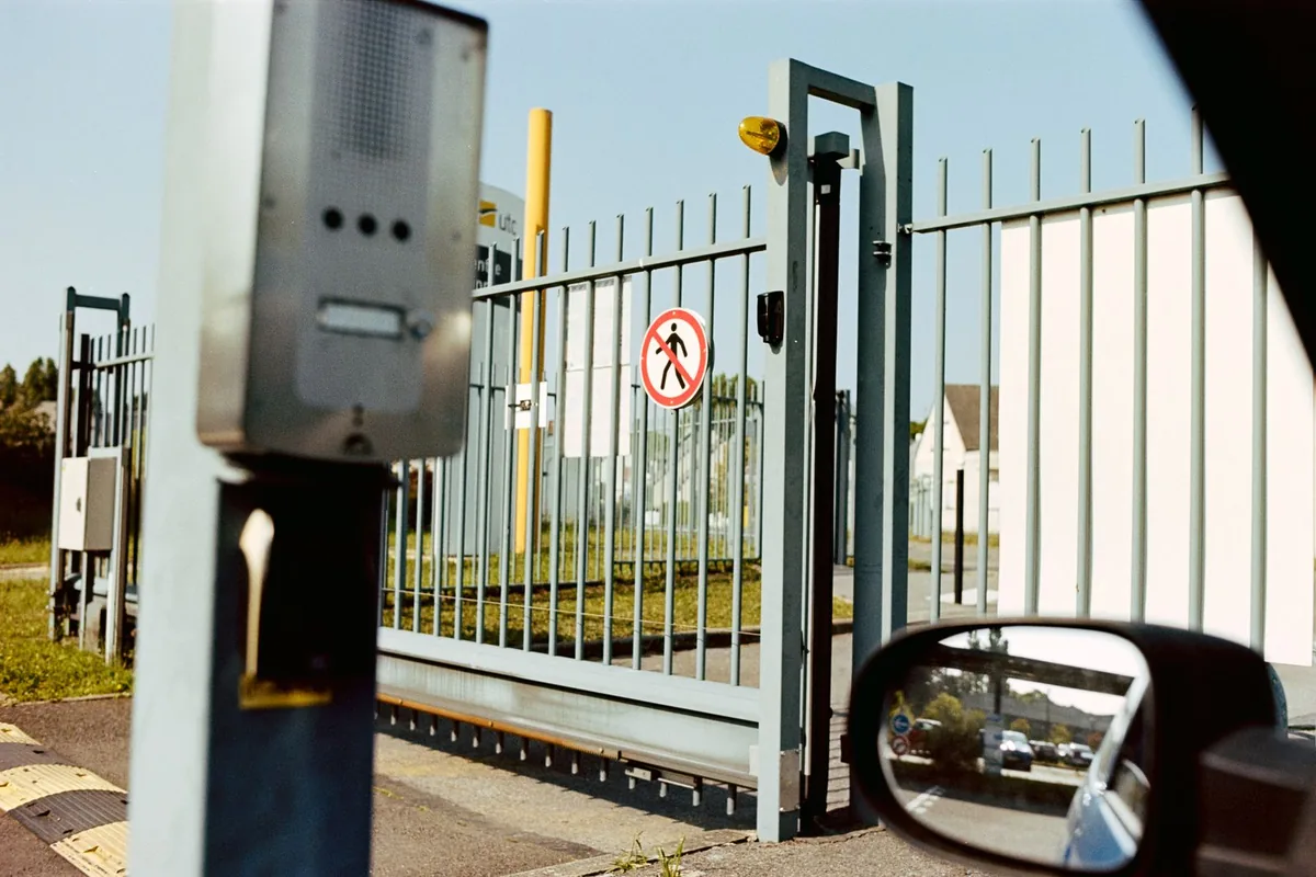 A closed sliding security gate with an intercom post beside it, seen from the driver's window of a waiting vehicle.