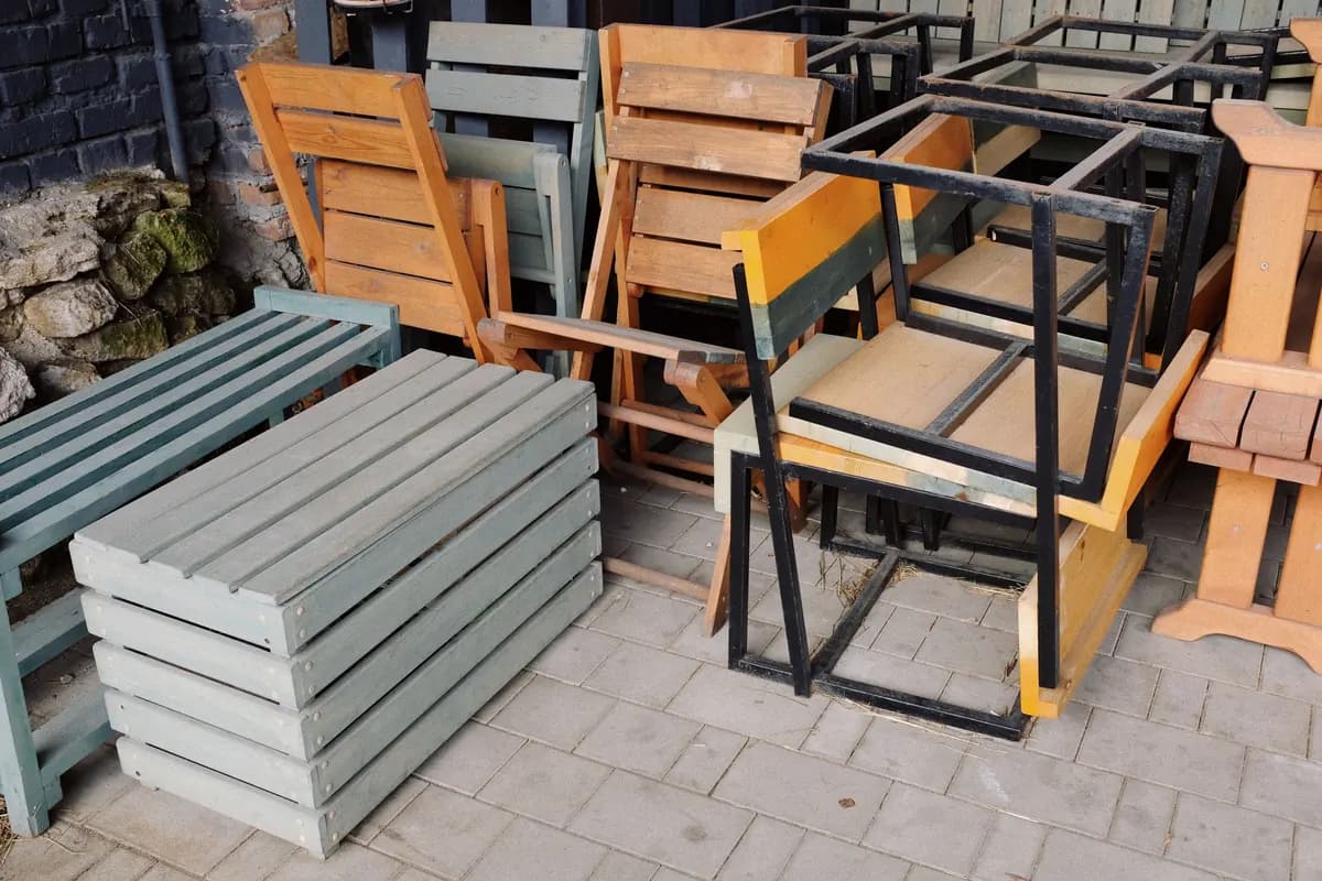 Wooden benches, folding chairs and stacked timber frames set aside together on a paved area beside a wall.