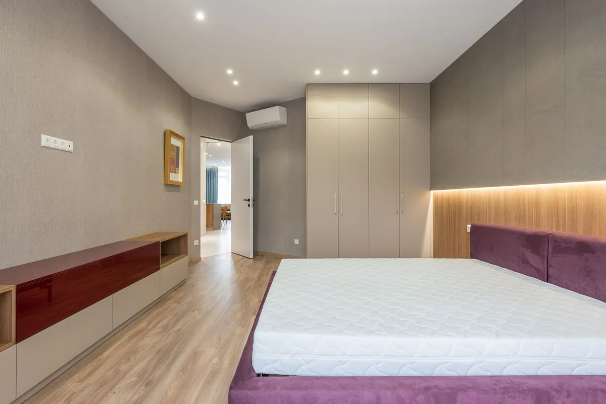 A stripped bed with a bare mattress on an upholstered base in a bedroom with fitted wardrobes along the far wall.