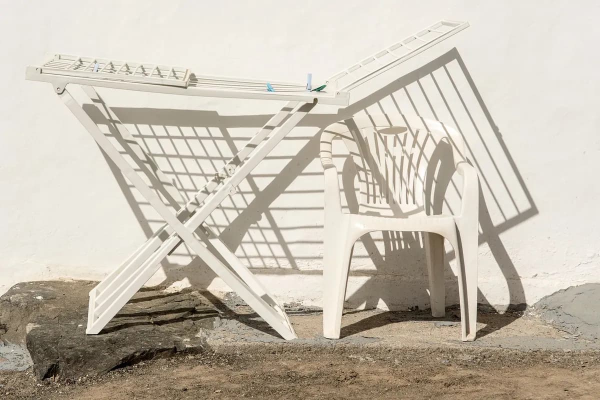A white folding plastic clothes-drying rack and a white plastic chair standing against a sunlit white wall, casting hard shadows.