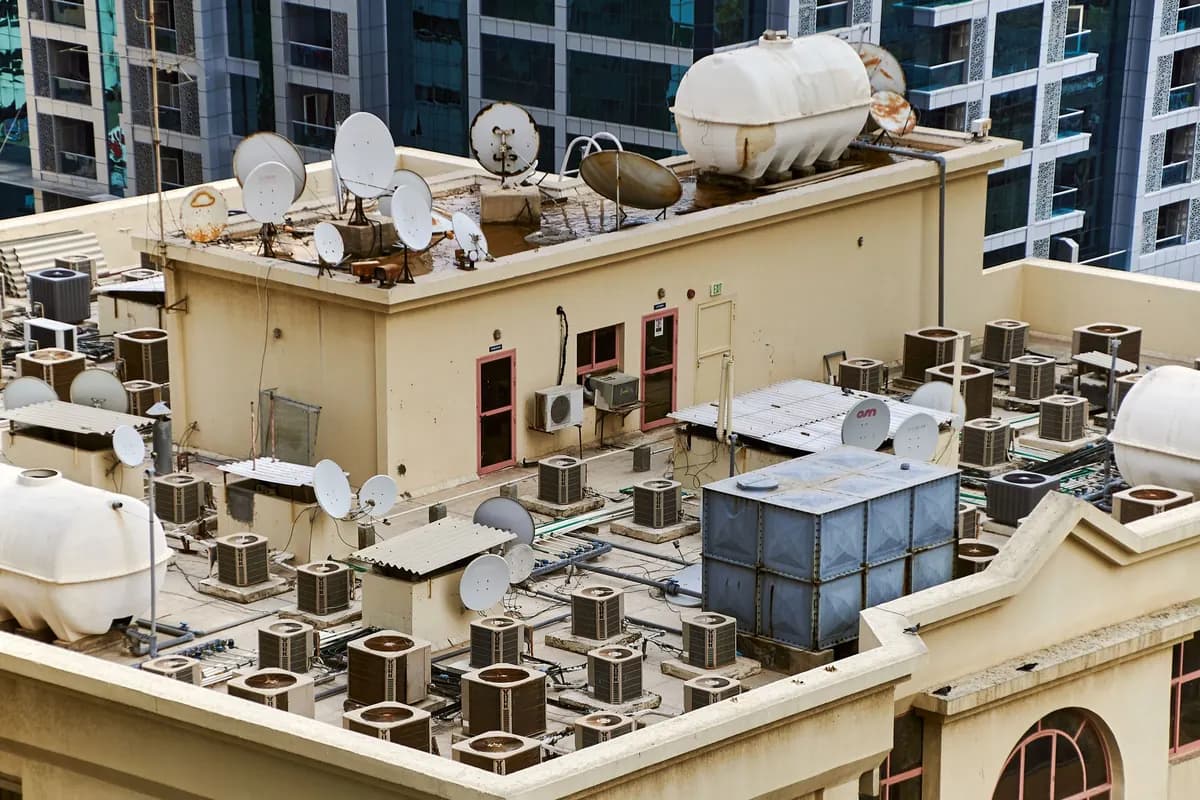 The flat roof of a residential tower covered with rows of air-conditioning condenser units, water tanks and satellite dishes around a plant room door.
