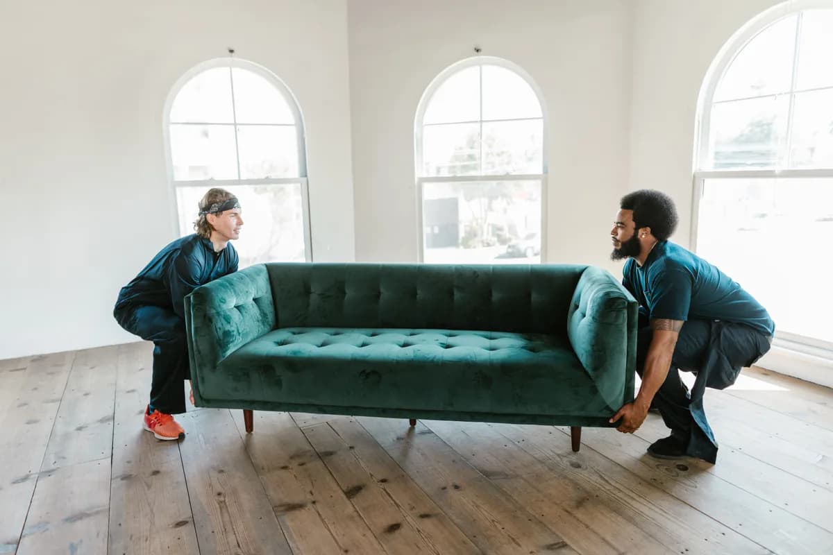 Two crew members crouched on either end of a sofa, lifting it together in an empty room.