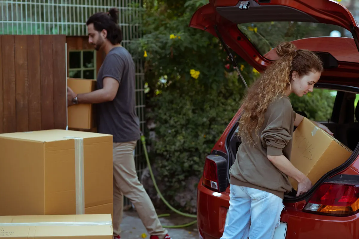 A woman lifting a labelled cardboard box into a red hatchback boot while a man carries another box past a garden fence.