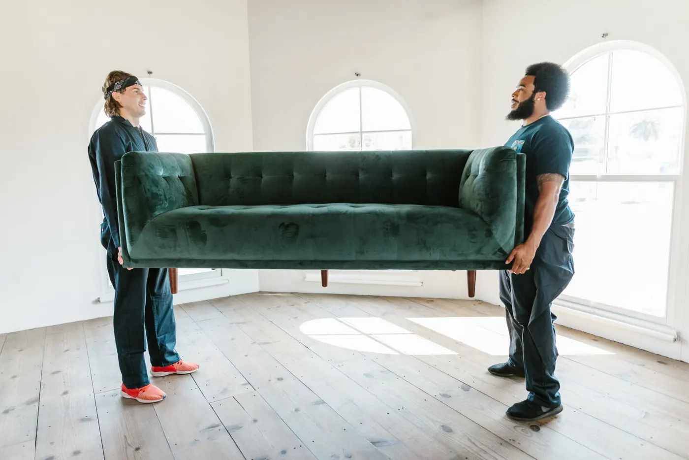 Two workers in dark uniforms carrying a green velvet sofa across an empty room with tall arched windows.