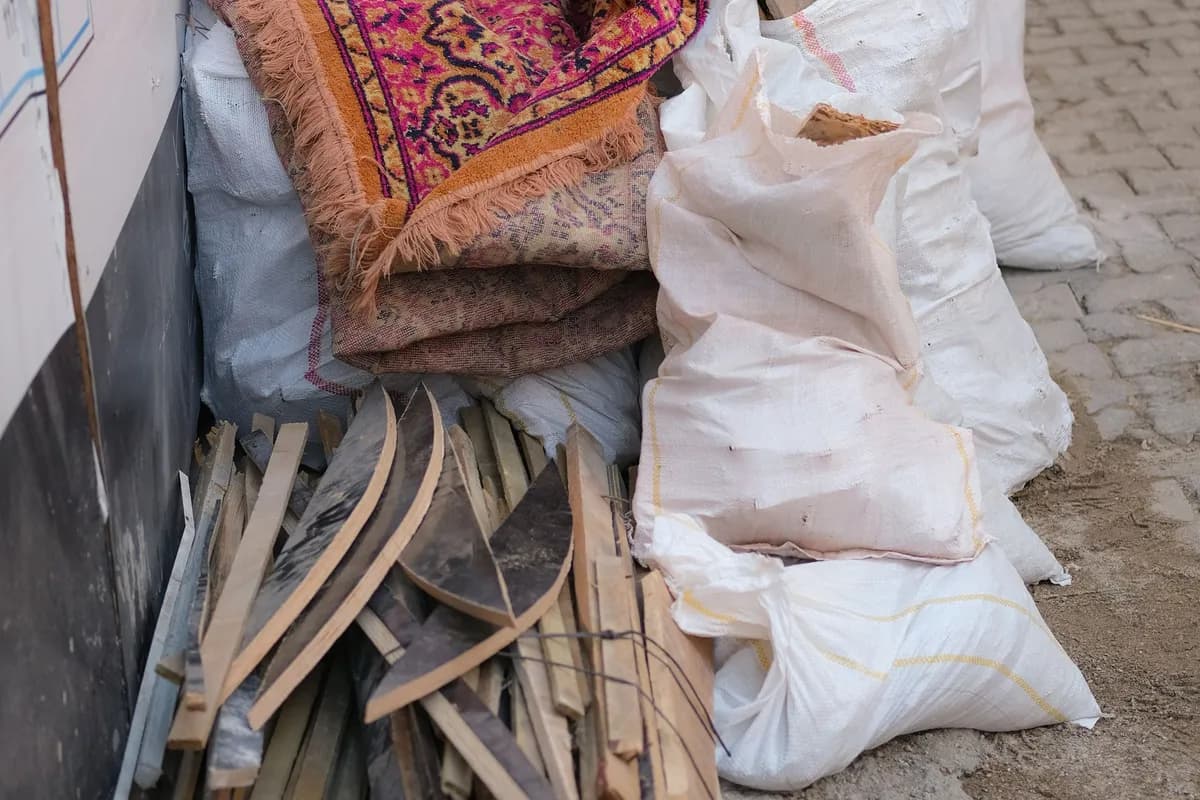A pile of rolled-up old carpets, timber offcuts and tied woven rubble sacks stacked against a wall on a pavement, waiting to be collected.