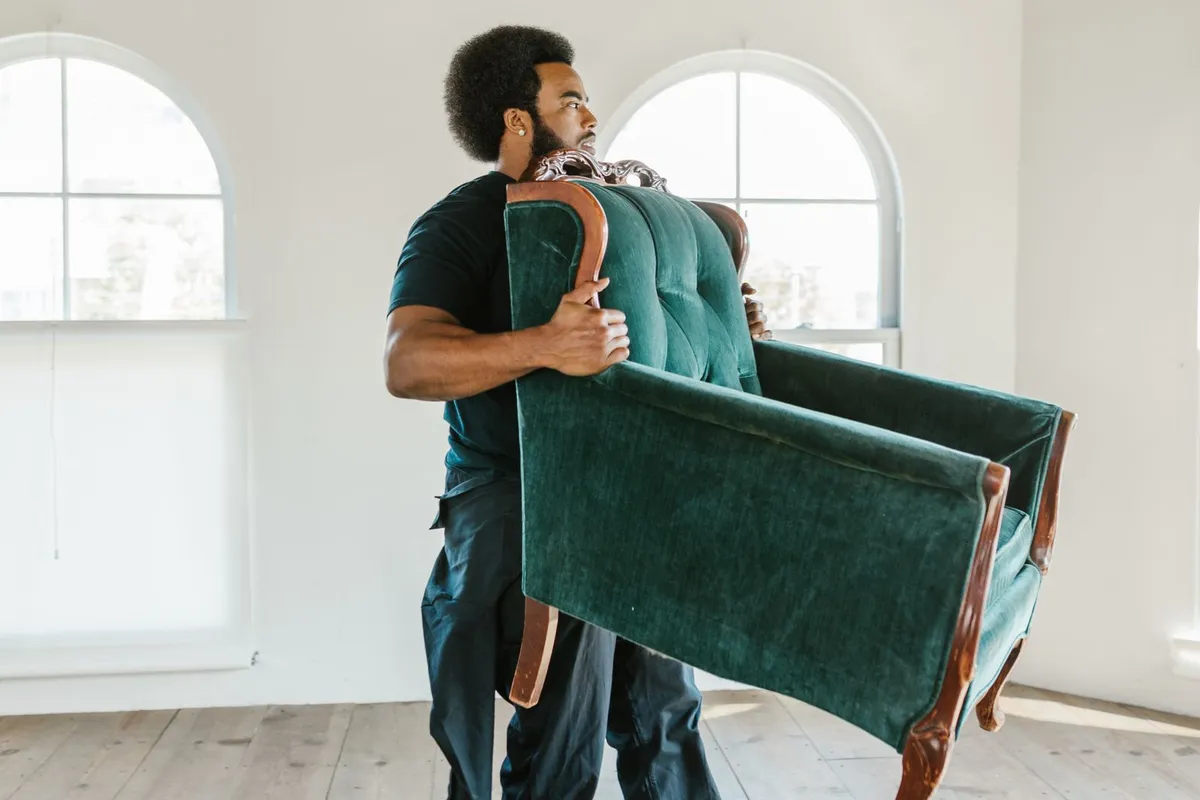 A man carrying a green upholstered armchair on his shoulder across a bright, empty room with bare floorboards and arched windows.
