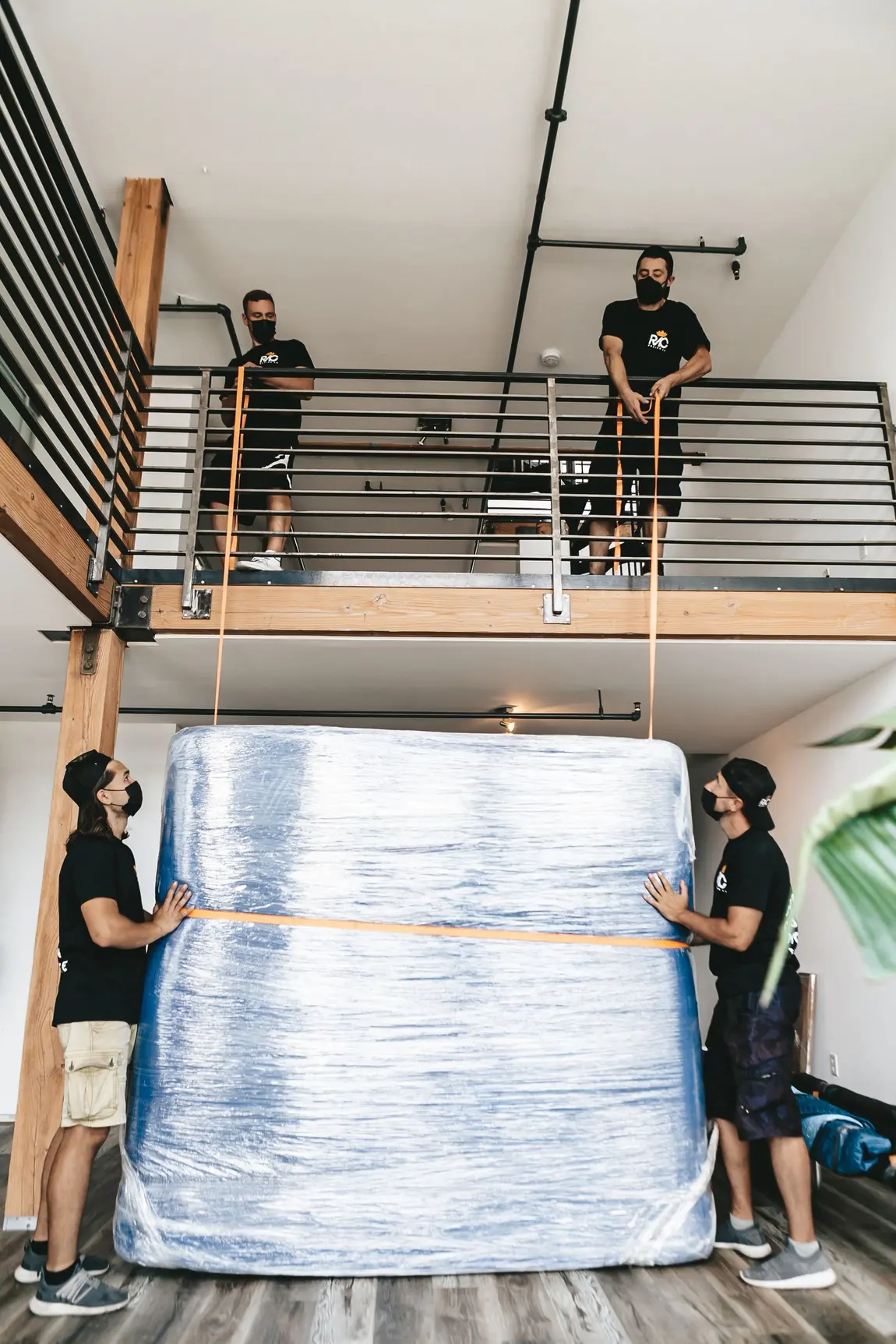 Four workers in black t-shirts using orange lifting straps to manoeuvre a large plastic-wrapped mattress inside a home with a mezzanine above.