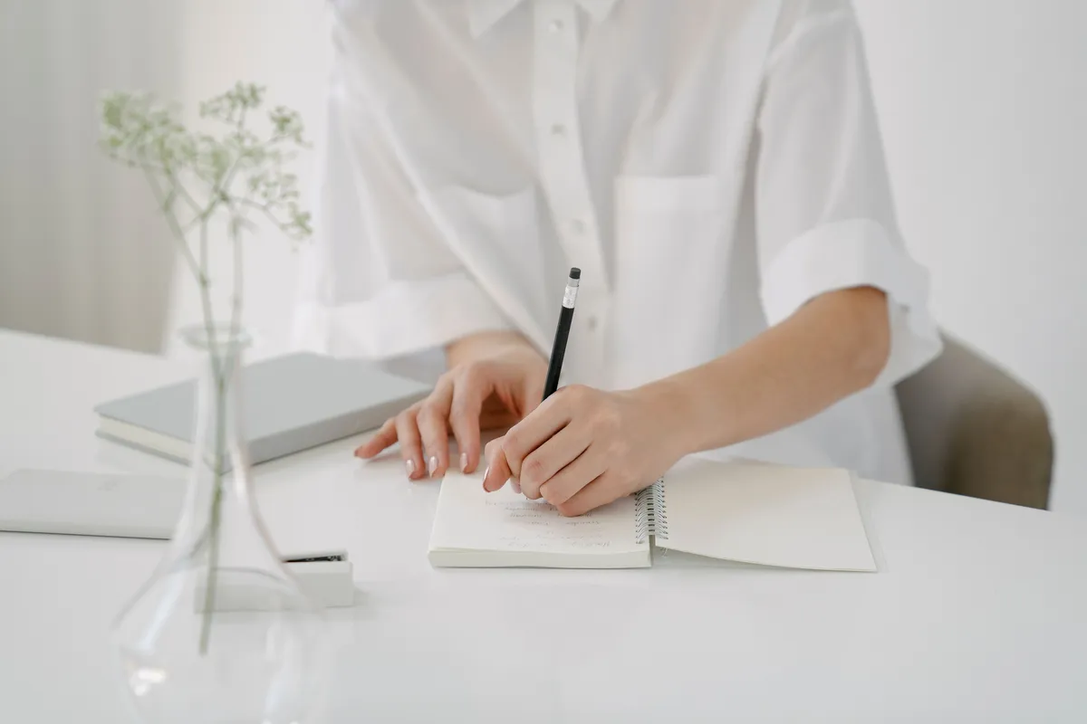 A person in a white shirt writing in a spiral notebook at a clean, minimalist desk.