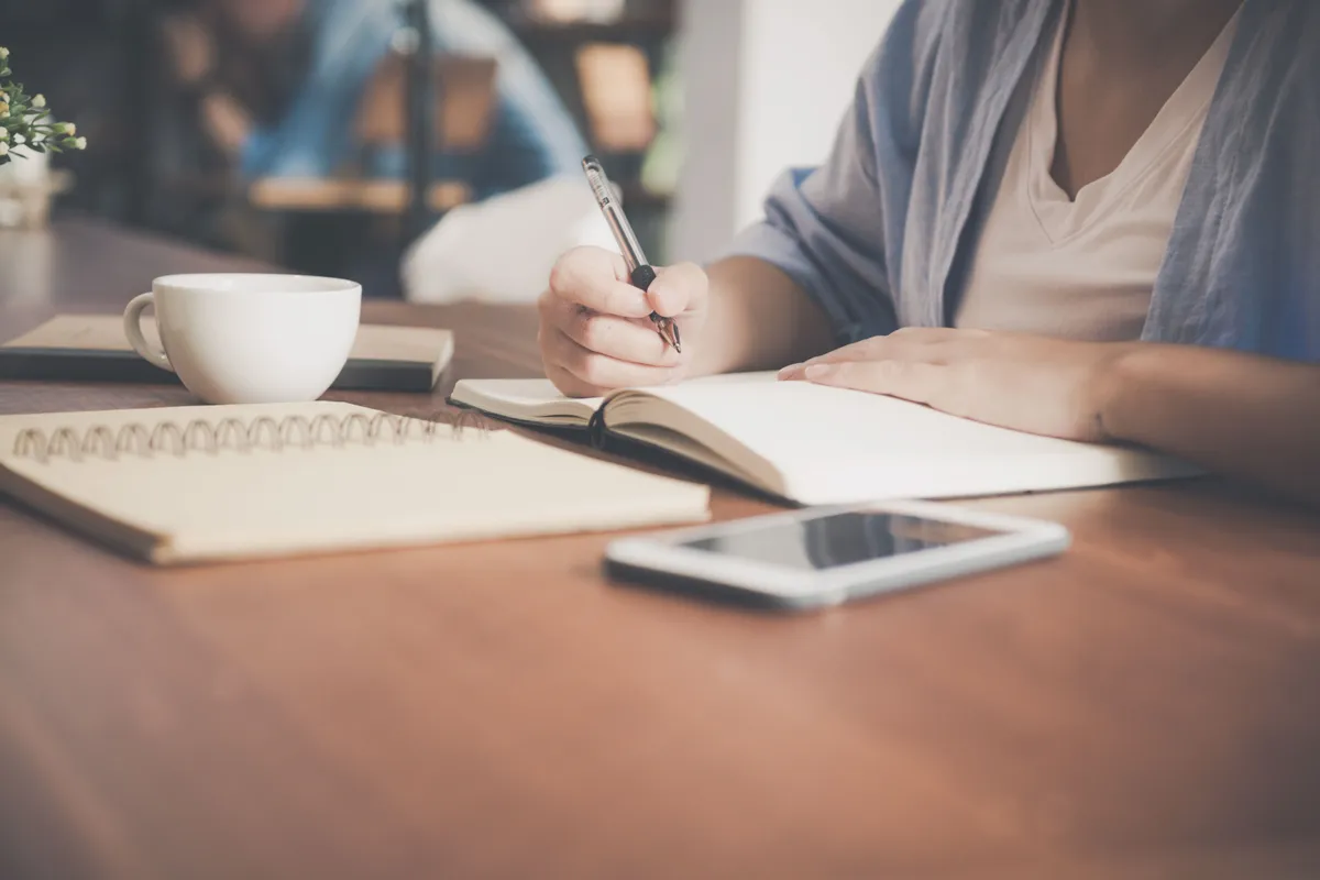 A hand writing in a notebook next to a smartphone and a cup of coffee on a wooden table.