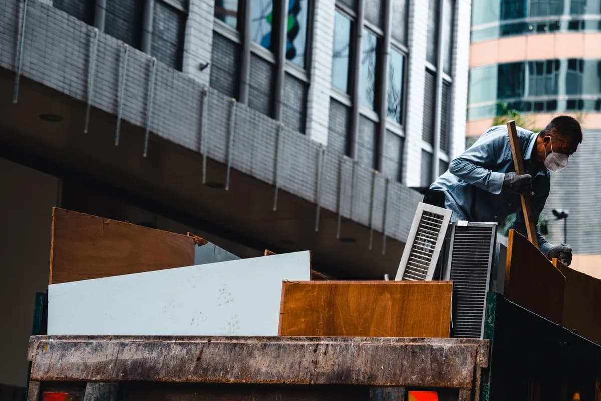 A worker in a face mask standing on an open-bed truck, loading large timber boards and metal grilles, with city office towers behind him.
