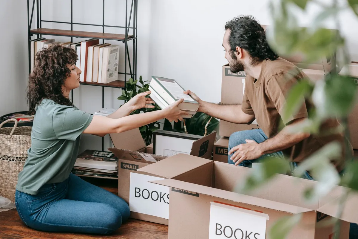 A woman passing a book to a man as they sort belongings into labelled cardboard boxes on the floor beside a bookshelf.