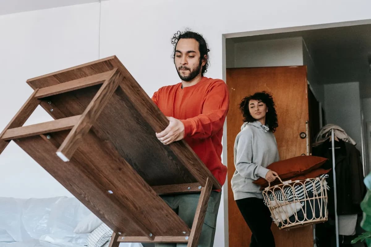 A man tilting a wooden shelf unit while a woman carries a basket of cushions towards the door of a bedroom with the bed covered in protective sheeting.