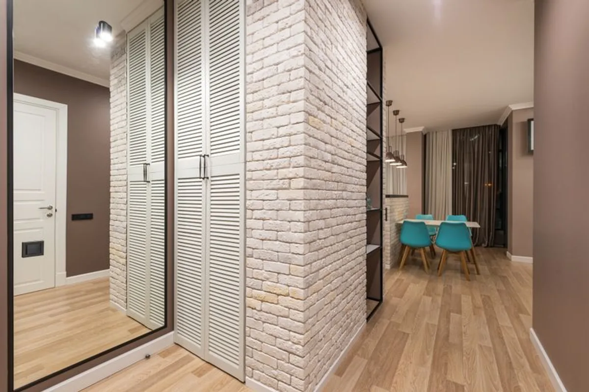 A modern apartment corridor turning past a brick corner pillar and fitted wardrobe, opening onto a dining area with wood flooring.