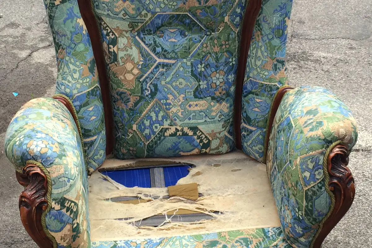 The seat of a patterned armchair worn and torn right through the covering, exposing the blue webbing strips and bare frame underneath.