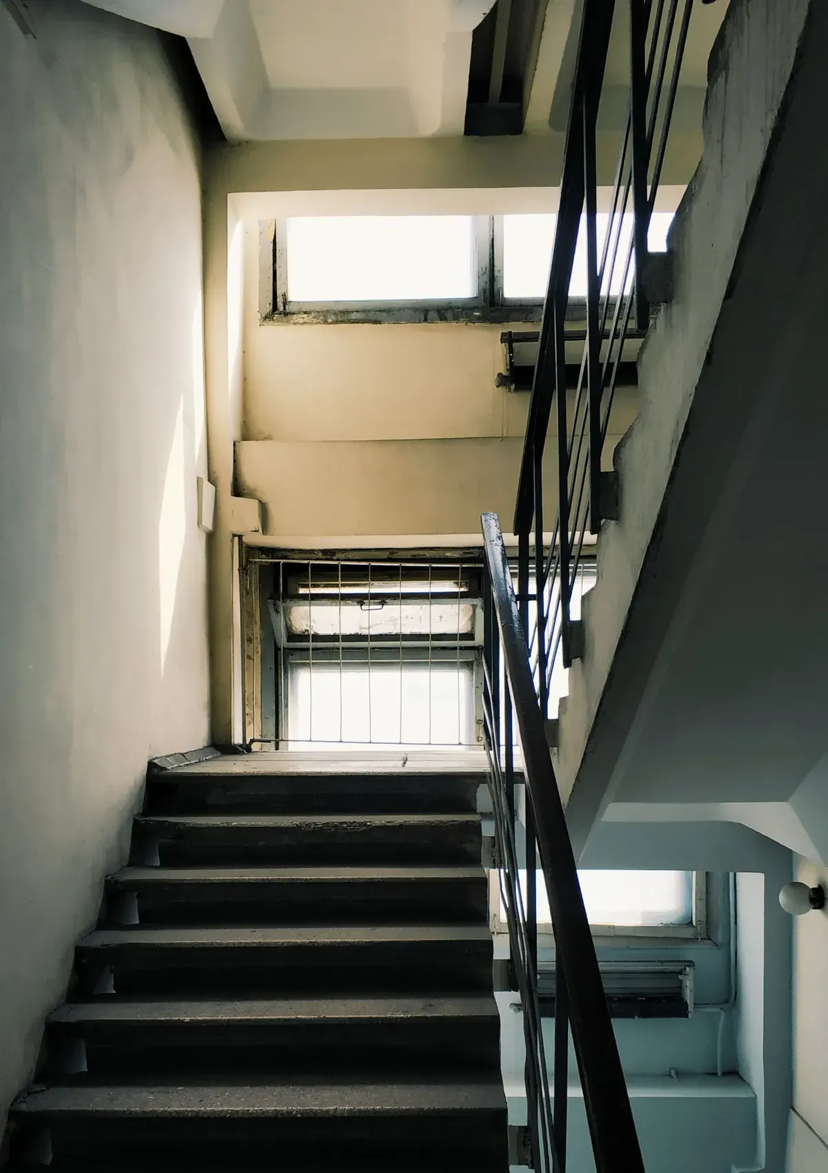 A concrete stairwell in a residential building, with a flight of steps rising to a half-landing beneath a window and a metal handrail along the open side.