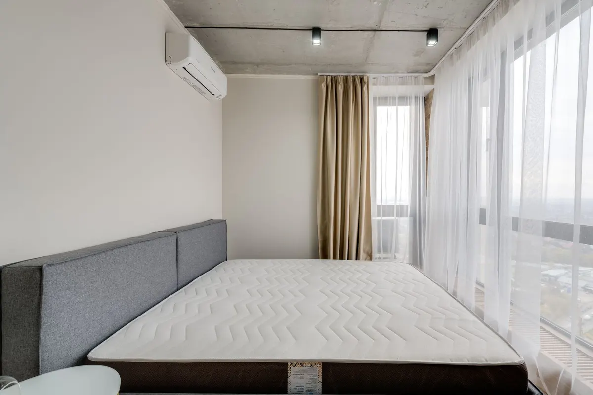 A stripped bare mattress on an upholstered bed base in a bright bedroom with sheer curtains and a wall-mounted air conditioning unit.