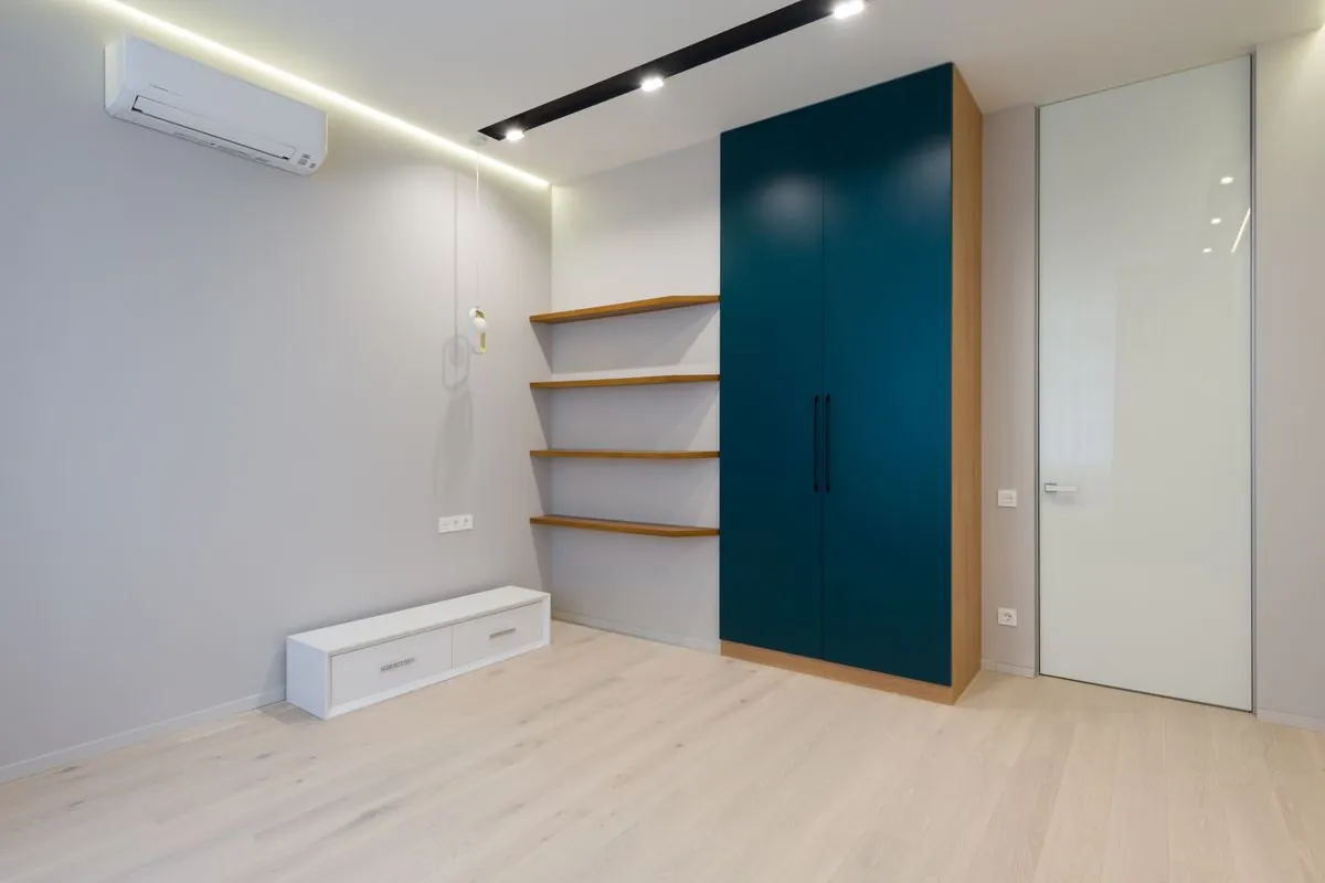 A tall two-door wardrobe, a low white drawer unit and a run of floating shelves standing in an otherwise empty modern apartment room.
