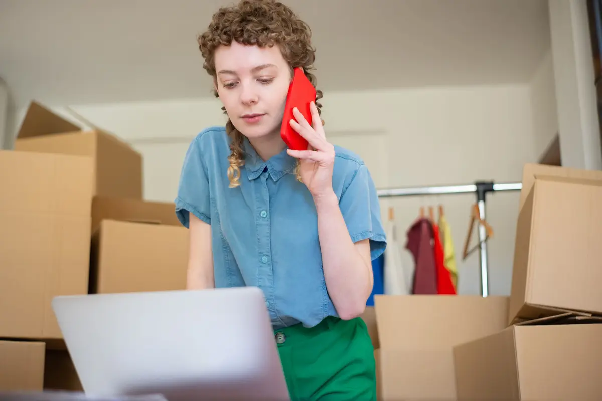 A woman standing among stacked cardboard boxes, speaking on a mobile phone while looking at an open laptop.