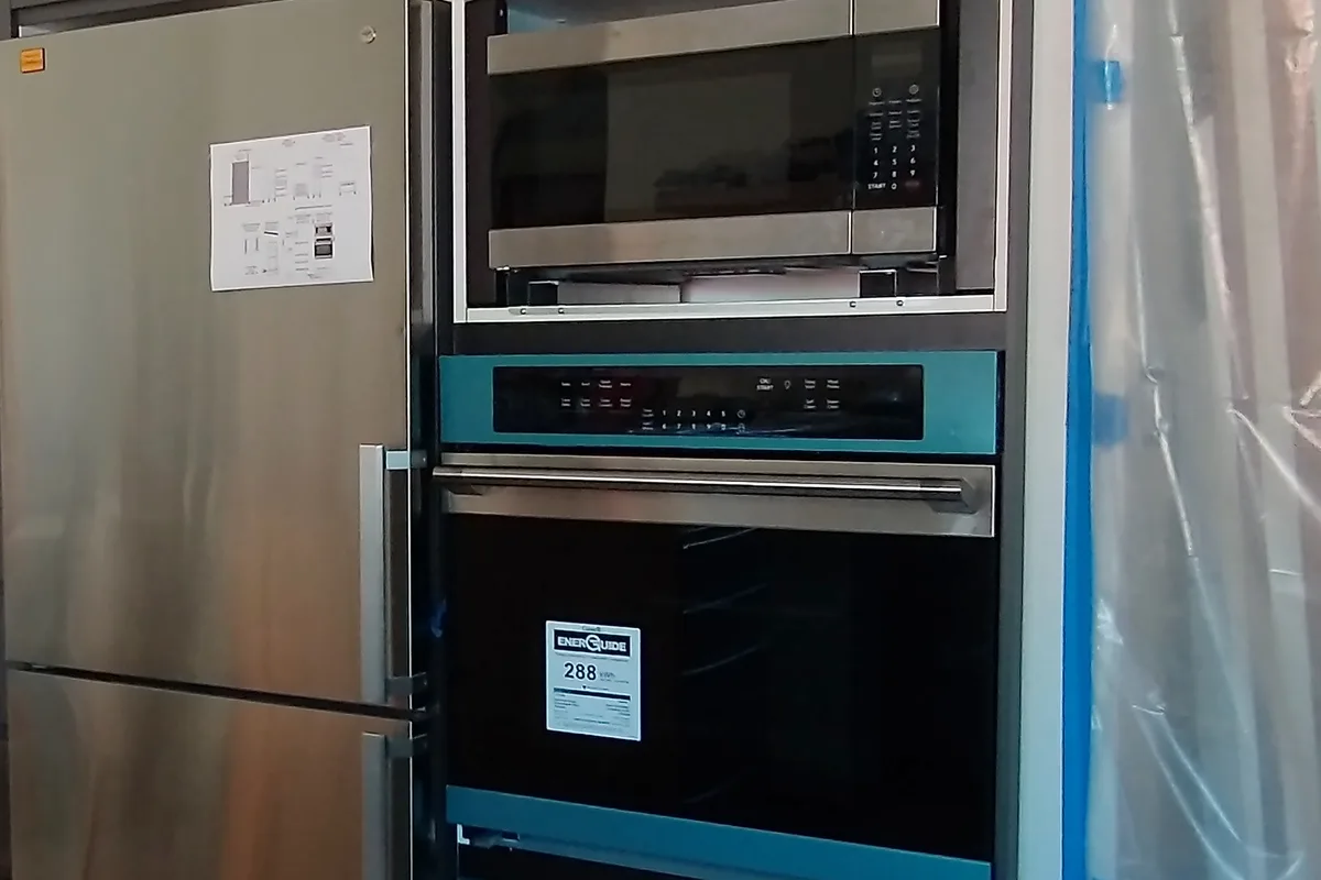 A built-in oven and microwave fitted into a tall kitchen cabinet beside a fridge, still covered in protective film.