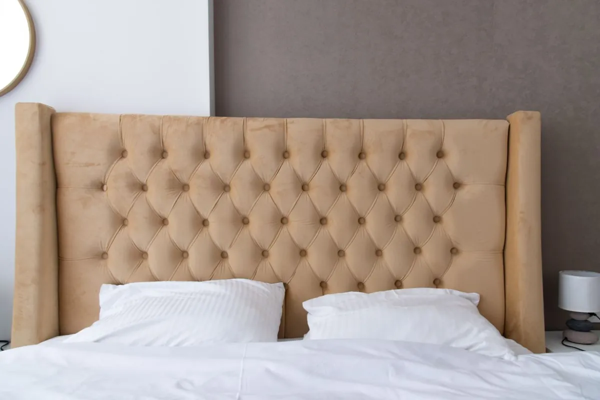 A tall buttoned fabric headboard with deep tufted folds and padded side wings standing behind two pillows on a made double bed.