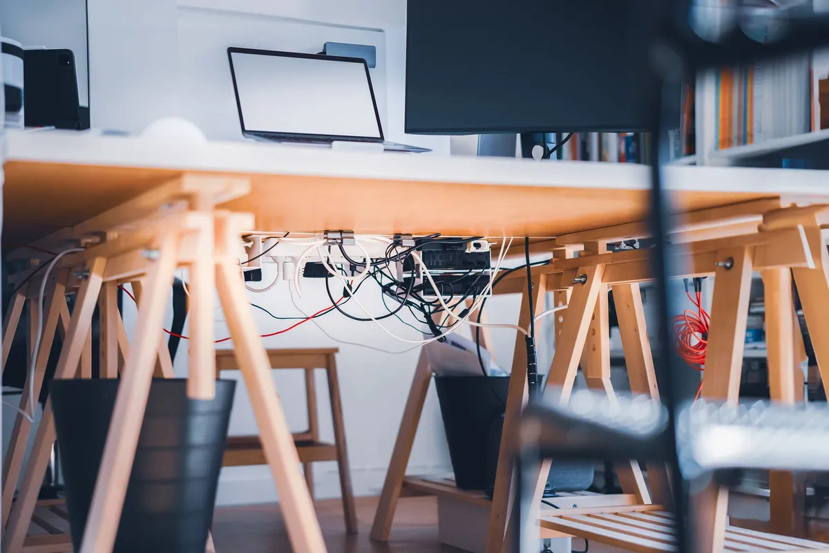 The underside of a home desk with power strips, a router and a tangle of black, white and red cables clipped to a tray beneath the top.