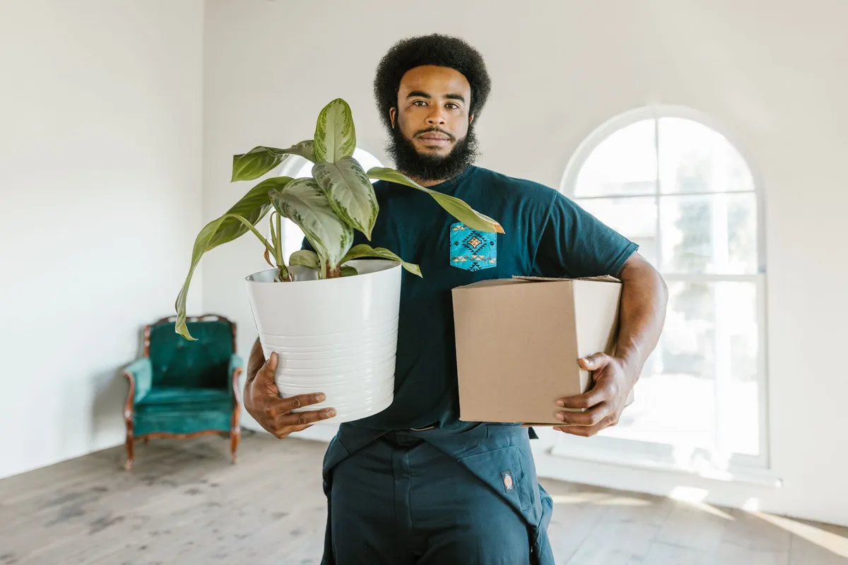 A worker in dark overalls carrying a potted houseplant and a cardboard box out of a bright, almost empty room.