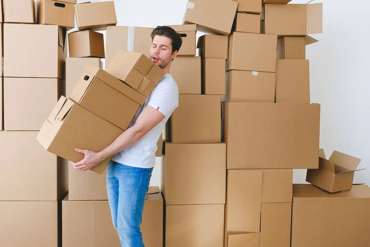 A man carrying three stacked cardboard boxes in front of a wall of piled cartons