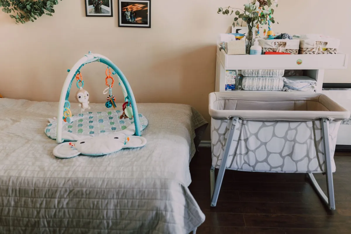 A white changing table stacked with folded linen and baskets beside a grey travel cot on a metal frame, with a padded play mat on the bed alongside.